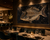 A large framed wall art piece depicting a fish made of sparkling gold-like stones against a dark starry background, displayed in a dimly lit restaurant interior with tables, chairs, and bar seating.