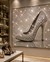 A large framed wall art piece featuring a single high-heeled shoe covered in sparkling rhinestones, displayed in a luxury shoe boutique with illuminated shelves and other heels around it.