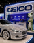 A luxury sedan covered in sparkling rhinestones and decorated with large GEICO logos, displayed on a platform at an auto show. A large GEICO sign, a green gecko statue, and a booth are visible in the background.