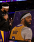 A large framed diamond painting portrait of basketball player, held by a man in a black suit while another man in a Los Angeles Lakers uniform stands beside it on a basketball court.